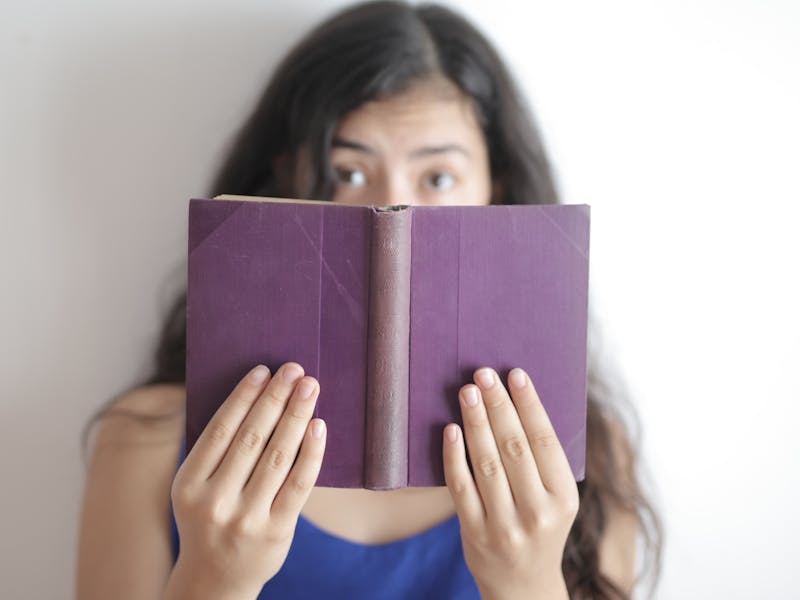 Young woman hiding face behind a purple book, curious eyes peeking.